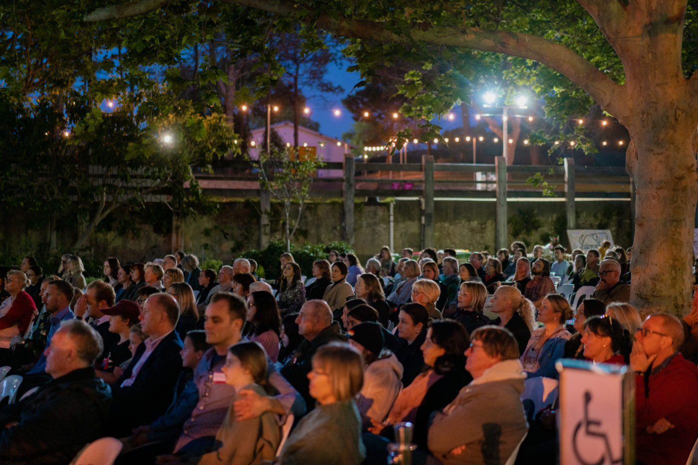 Crowd shot of people sitting down at night under a tree with fairy lights around them