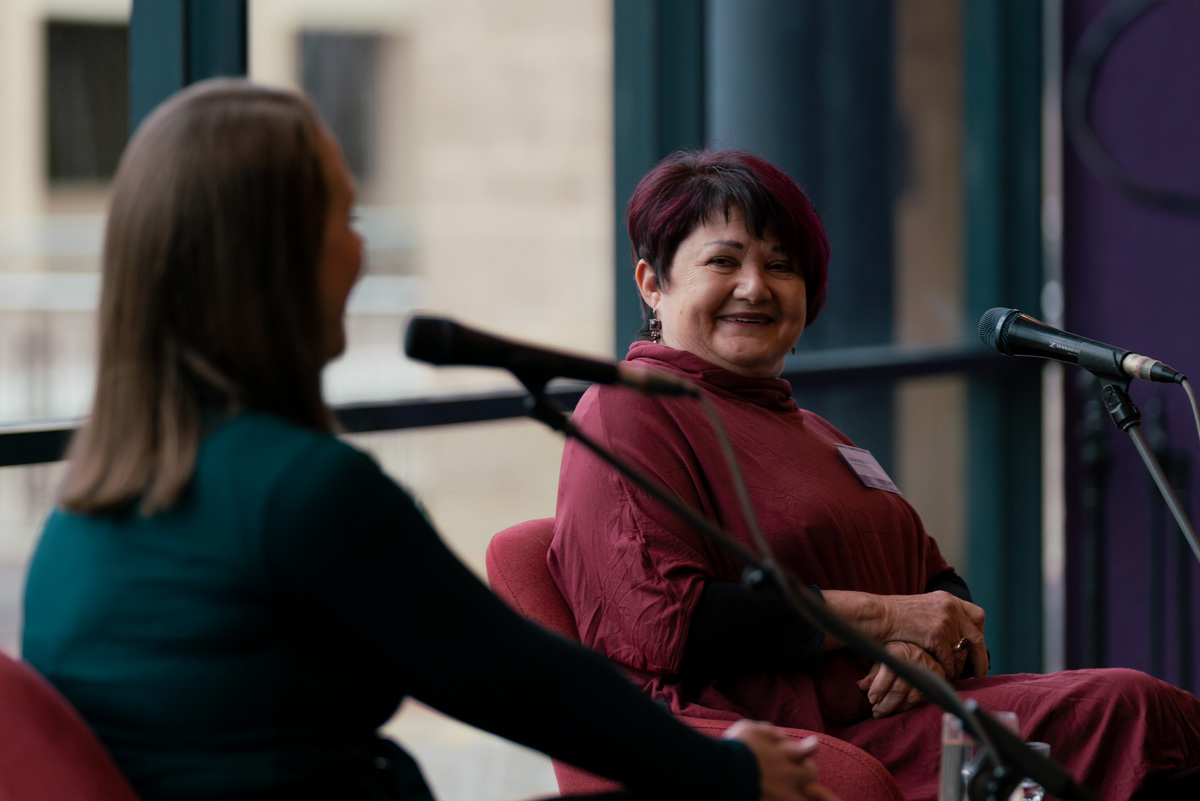 Image of a woman seated in front a microphone smiling at another woman also seated 