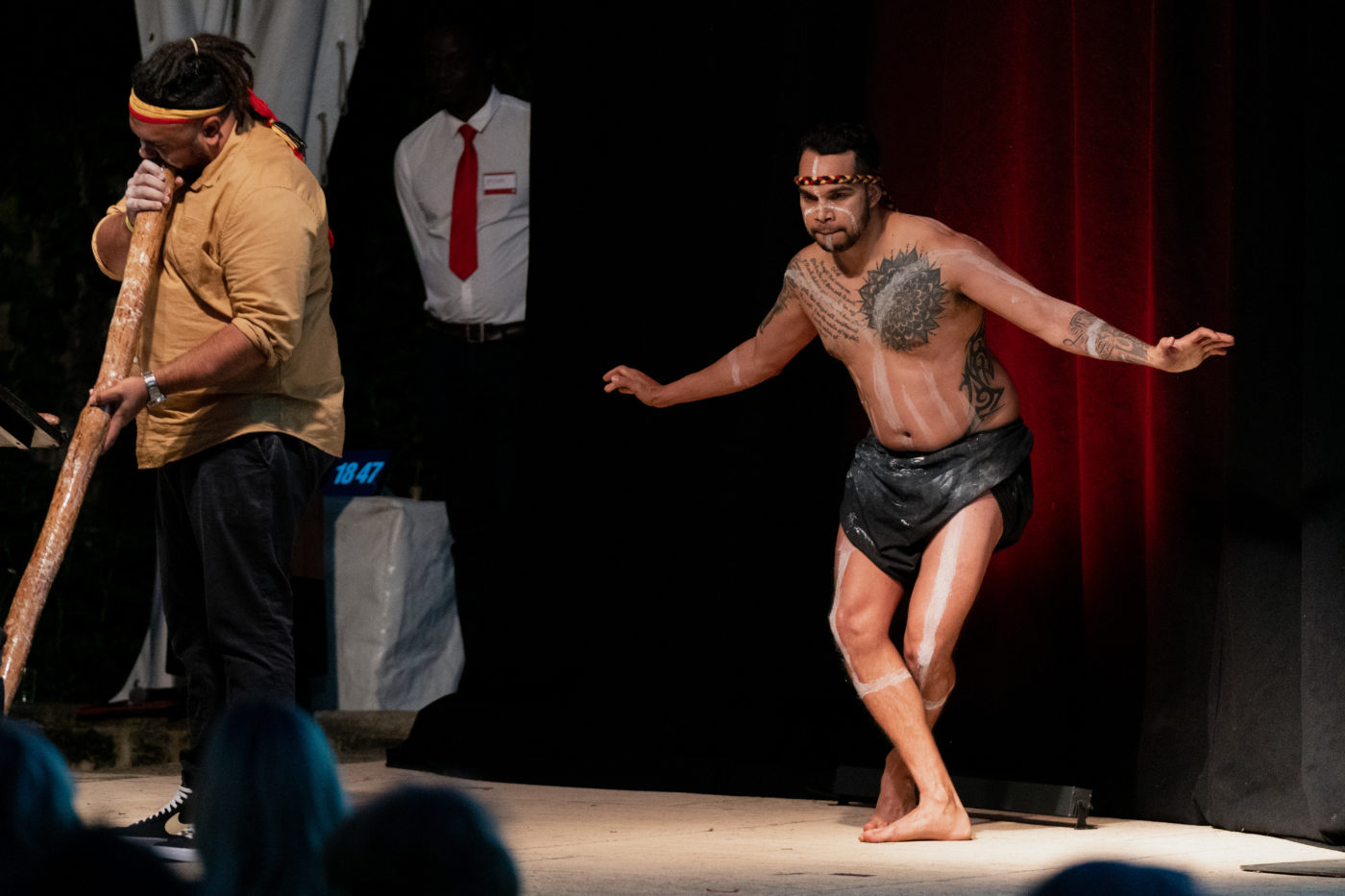 Two men, one playing the didgeridoo and the other in a dance position.