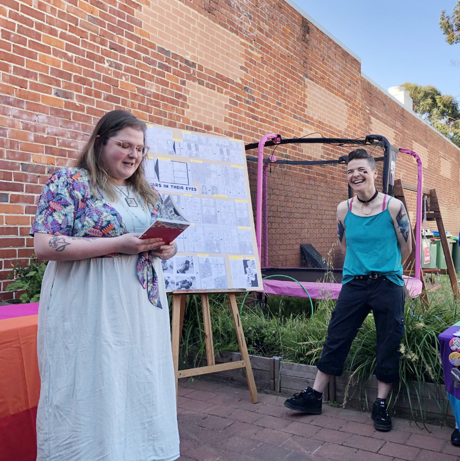 Image of two women, one in a white dress holding a book and the other standing to the side in front of an easel