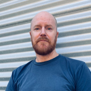 Image of a man in a blue shirt leaning on a grey garage door.